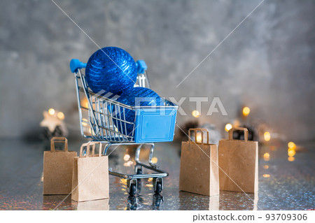 Holiday shopping. Shopping cart with blue shiny balls and paper bags on a gray background with garland and lights. Christmas concept 93709306