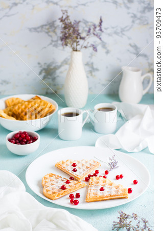 Heart shaped Belgian waffles with red berries and powdered sugar on a plate and two cups of coffee on the table. Valentine's Day. Vertical view 93709473