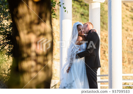 Young beautiful interracial newlyweds hugging against the background of white columns Young beautiful interracial newlyweds hugging against the background of white columns 93709583