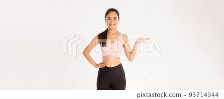 Sport, wellbeing and active lifestyle concept. Portrait of cheerful attractive asian fitness coach, female athlete introduce or demonstrate product for workout, holding something on hand 93714344