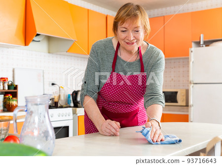 Elderly woman housewife in apron cleaning furniture at kitchen 93716643