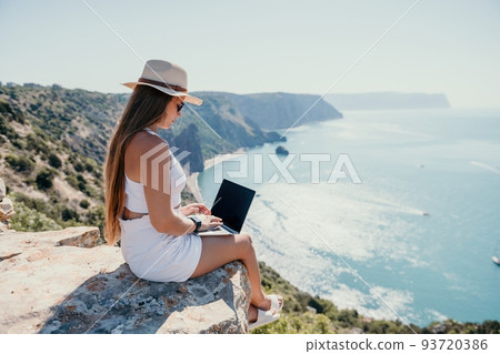 Digital nomad, woman in the hat, a business woman with a laptop sits on the rocks by the sea during sunset, makes a business transaction online from a distance. Freelance, remote work on vacation. 93720386