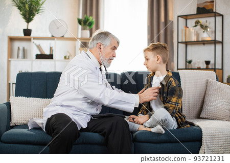 Boy sitting on the couch while caring experienced nice mature male doctor listening his heartbeat 93721231