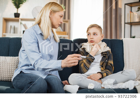 Mother taking care of her cute sick teen boy checking temperature, sitting on the sofa. Mother taking care of her cute sick teen boy checking temperature, sitting on the sofa. 93722306