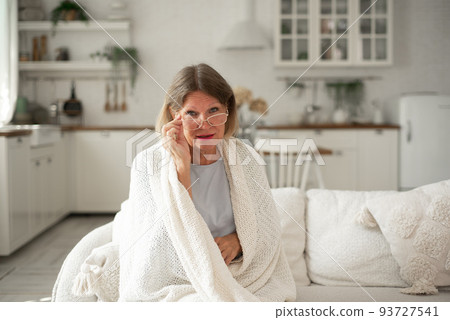 An elderly woman in glasses in a plaid is warming herself on the sofa at home. 93727541