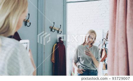 Pretty blond woman is making mirror selfie with smart phone while standing in nice fitting room in luxurious clothes boutique. She is posing, moving and smiling carelessly. 93729007