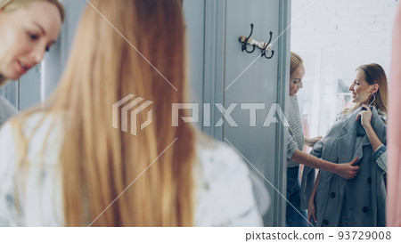 Pretty young woman is checking fashionable coat in fitting room with her friend helping her to appraise garment. They are talking, gesturing and looking at clothing in large mirror. Pretty young woman is checking fashionable coat in fitting room with her friend helping her to appraise garment. They are talking, gesturing and looking at clothing in large mirror. 93729008