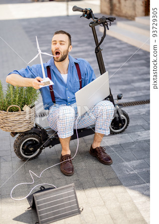 Man blows on toy wind generator while sitting with laptop and green plant on electric scooter outdoors 93729305