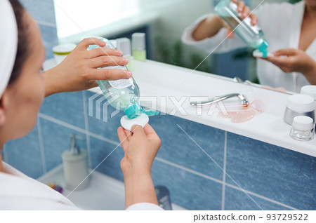 Selective focus on the hands of a blurred woman standing in front of a bathroom mirror and pouring a cleansing lotion on a cotton pad, to clean the face skin and remove make-up. 93729522