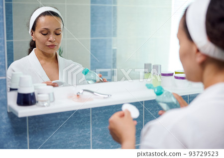 Reflection in bathroom mirror of a dark-haired African American young pretty woman, wearing white bathrobe, using a cotton pad and a face cleanser to remove make-up. Healthy skin is a daily commitment 93729523