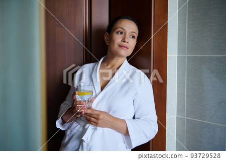 Charming confident middle aged woman in a white bathrobe with a glass of refreshing lemon water for a good start to the day. Detox care for healthy skin and body. Anti-aging, healthy lifestyle concept 93729528