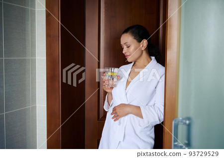 Charming relaxed brunette woman in a white waffle robe, holding a glass of lemon water while standing th home bathroom. Detox care for healthy skin. Body care. Healthy lifestyle. Morning routine 93729529