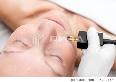 Close-up beautician doctor hand making anti-age face remodel procedure treatment with hardware microcurrent electric tool attractive young 30-40 female model at beauty clinic. Cosmetologist skincare 93729532