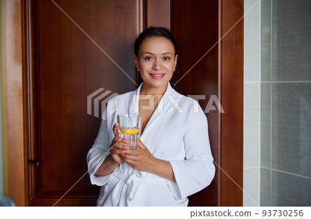 Close-up portrait. Beautiful attractive Hispanic woman with glowing skin, in bathrobe, smiles looking at camera, holds glass with lemon water for good start to the day. Diet, vitality, healthy habits 93730256