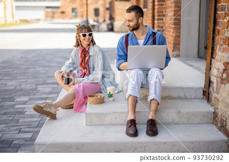 Stylish colleagues work on laptop and eat take away food on a street outdoors 93730292