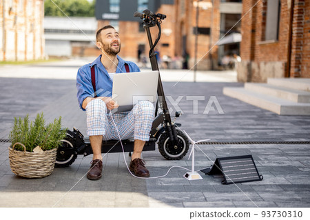 Man charges laptop with solar panels while sitting on electric scooter outdoors 93730310
