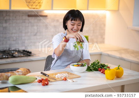 A young asian woman in the kitchen cooking and looking contented A young asian woman in the kitchen cooking and looking contented 93733157