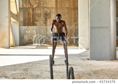 Rope workout. Sport man doing battle ropes exercise outdoor. Black male athlete exercising, doing functional fitness training with heavy rope Rope workout. Sport man doing battle ropes exercise outdoor. Black male athlete exercising, doing functional fitness training with heavy rope 93734379