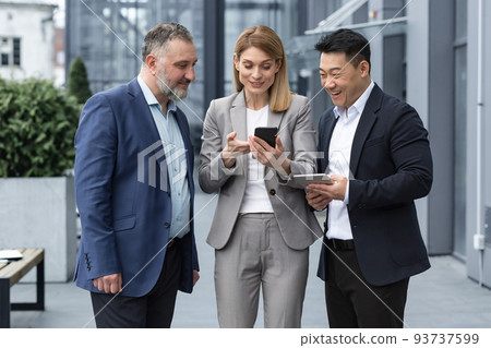 three IT specialists entrepreneurs outside office building watching video on colleague's phone, happy and smiling resting on break from work 93737599