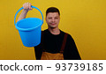 A young guy gardener or cleaner in uniform holds a blue bucket in his hands 93739185