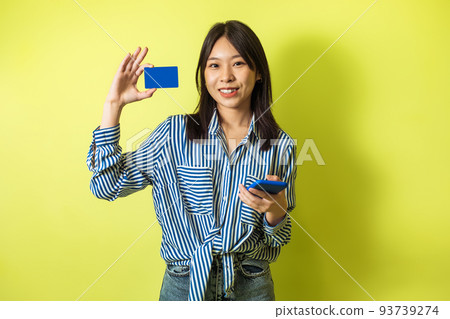 Korean Lady Shopping Via Phone Showing Credit Card, Yellow Background Korean Lady Shopping Via Phone Showing Credit Card, Yellow Background 93739274