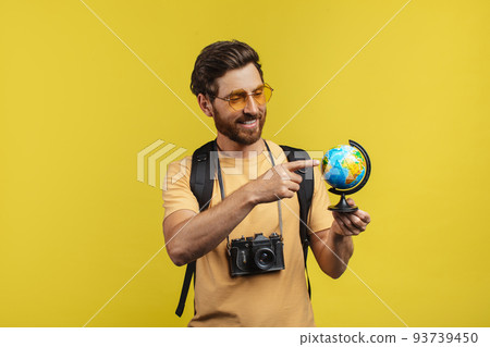 Happy male traveler holding world globe and pointing on it, wearing backpack and camera, yellow background Happy male traveler holding world globe and pointing on it, wearing backpack and camera, yellow background 93739450