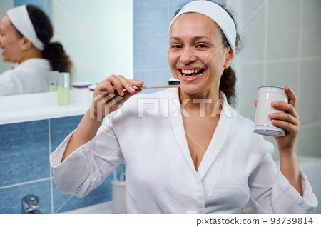 Multiethnic middle aged woman in white bathrobe, enjoys morning beauty procedures, brushing teeth with eco bamboo toothbrush and whitening toothpaste, smiles beautiful toothy smile, looking at camera 93739814