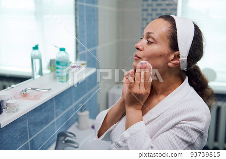 Close-up portrait of a natural beauty woman in white bathrobe, using cleanser cosmetic lotion and cotton pad, removes makeup, enjoys skin care procedures, looking at her reflection in bathroom mirror 93739815