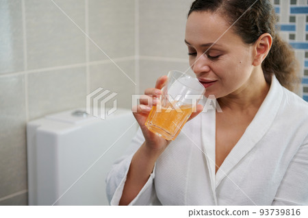 Multi-ethnic pretty woman in white bathrobe, drinking water with effervescent painkiller pills, according to physician's prescription. Woman suffering head ache. Migraine and fever, pain relievers. 93739816