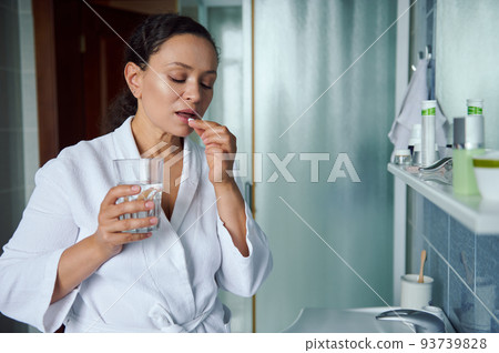 Middle-aged multi-ethnic woman in white bathrobe, taking daily dose of complex healthcare skin, hair and nails omega vitamins and drinking glass of fresh pure water. Immunity improvement concept. 93739828