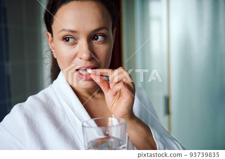 Headshot of multiethnic beautiful woman wearing white bath robe, taking medication or nutritional supplements for skin care, according to medical prescription. Antidepressant therapy. Painkiller pills 93739835