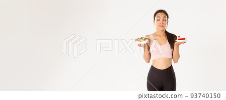 Active lifestyle, fitness and wellbeing concept. Indecisive cute asian slim girl in workout outfit, holding healthy salad and cake, hesitating, need stay on diet, losing weight, white background 93740150
