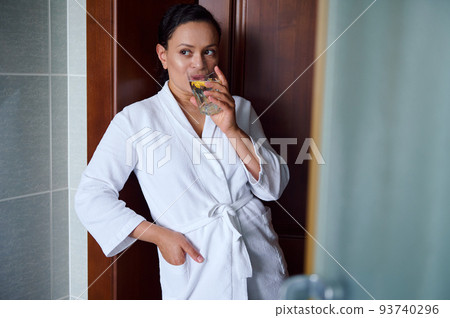 Beautiful African American woman in a white waffle bathrobe drinks refreshing lemon water for healthy and glowing skin. Detox treatment for a good start to the day. Healthy lifestyle. Body care 93740296