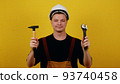 Friendly plumber foreman or handyman in hardhat holding adjustable wrench and hammer on yellow isolated background 93740458