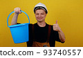 A young guy gardener or cleaner in uniform holds a blue bucket in his hands and points his finger at it 93740557