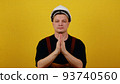 Young guy of caucasian appearance wearing a hard hat and builder clothes over isolated yellow background praying with his hands with a hopeful expression on his face and is very worried 93740560