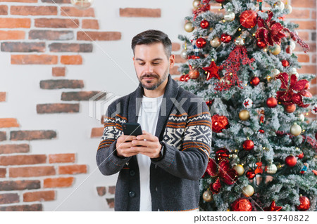 A serious and focused man is corresponding on a mobile phone near the Christmas tree at home A serious and focused man is corresponding on a mobile phone near the Christmas tree at home 93740822