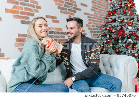 Young family together celebrate New Year and Christmas, couple man and woman give gifts to each other sitting on sofa at home happy and in love 93740870