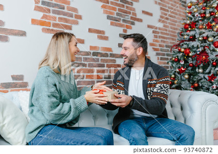 Young family together celebrate New Year and Christmas, couple man and woman give gifts to each other sitting on sofa at home happy and in love 93740872