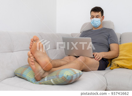 young man in mask and with laptop sitting on sofa 93741662