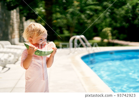 Little girl eats a piece of watermelon standing near the pool 93742923