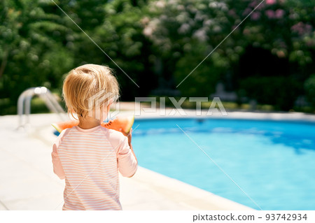 Little baby stands by the pool, holding a watermelon in his hands. Back view 93742934