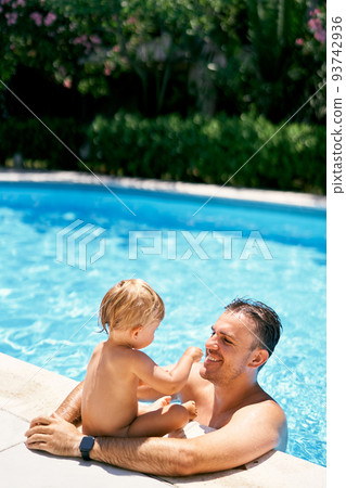 Smiling dad stands in the water and hugs a small child sitting on the edge of the pool 93742936