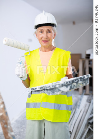 Portrait of elderly positive woman painter in protective helmet and yellow vest who paints walls with paint roller Portrait of elderly positive woman painter in protective helmet and yellow vest who paints walls with paint roller 93746846