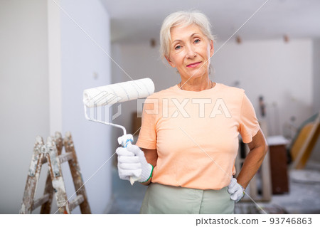Mature woman painting walls with paint roller in refurbished room Mature woman painting walls with paint roller in refurbished room 93746863