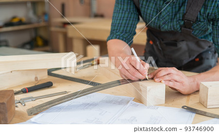 A carpenter measures wooden planks and makes marks with a pencil in a workshop. 93746900