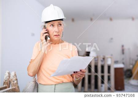 Elderly cottage owner in protective helmet talking on mobile phone in renovated room 93747108