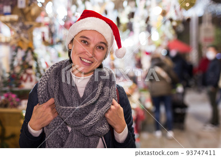 Smiling young woman in Santa hat standing at street Christmas fair Smiling young woman in Santa hat standing at street Christmas fair 93747411