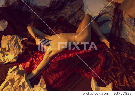 Conceptual portrait of young tender girl in lingerie lying on floor with red cloth, paper sheets over dark background. Mental health, art, creation 93767855