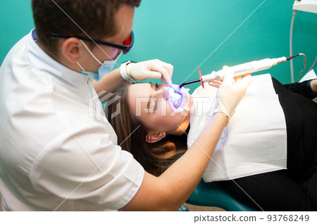 Dentist using whitening ultraviolet lamp on her teeth. Young girl patient at the dental clinic lies in uv glasses on the dentist's chair.. 93768249